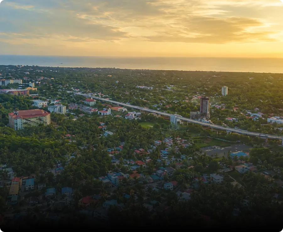 Aerial view of a city with abundant greenery, scattered buildings, and a highway running through it at sunset, with the sky glowing orange above the horizon.